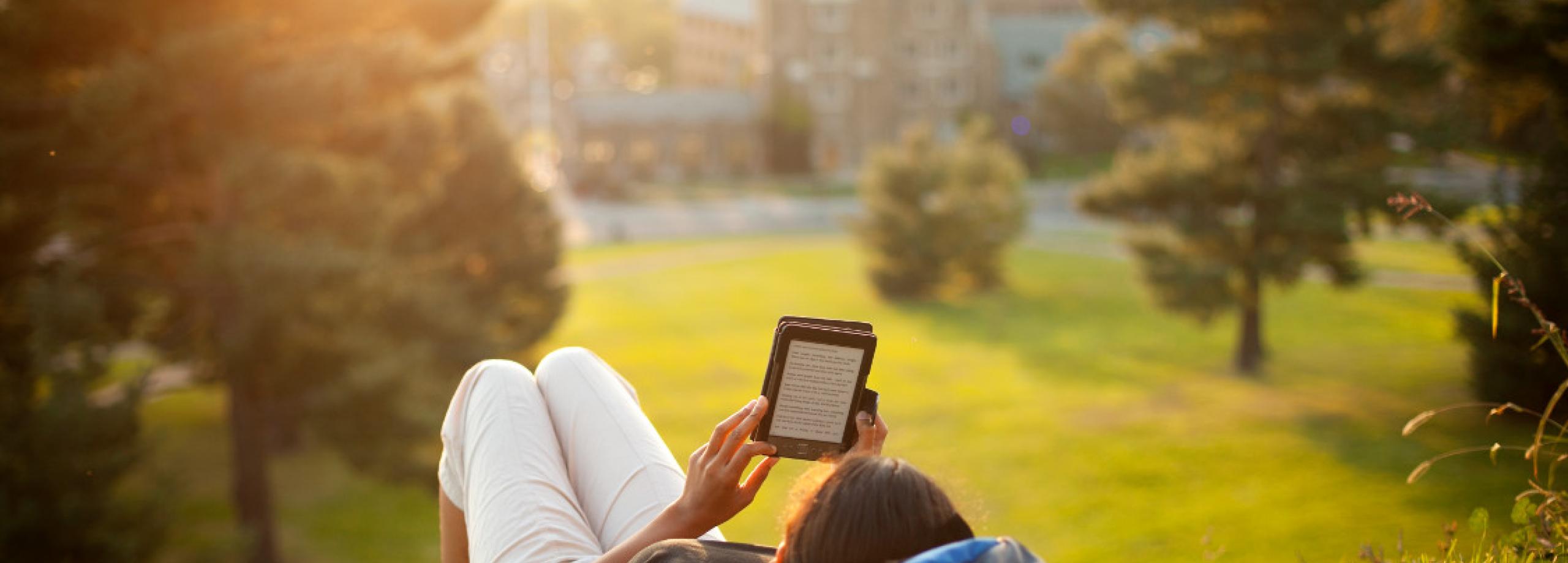 A student laying on a hill on campus, reading from a tablet